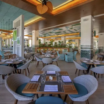 salle de restaurant, chaises confortables grises, tables en bois carrées, luminaires bas, piliers carrés blanc et verts, décoration volets blancs verts et bleus au plafond