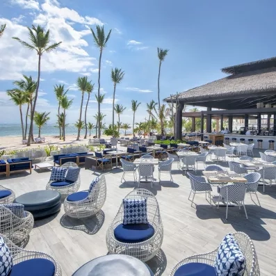 terrasse restaurant extérieur, fauteuils et coussins bleus et blancs, pieds dans l'eau, toit du bar paillote, palmiers et plage de sable, océan bleu et ciel bleu avec quelques nuages