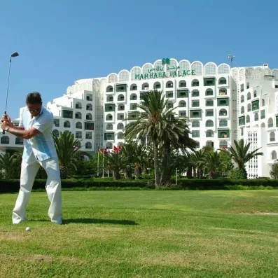 Homme jouant au golf sur le green verdoyant devant la façade blanche de l'hôtel Marhaba Palace.