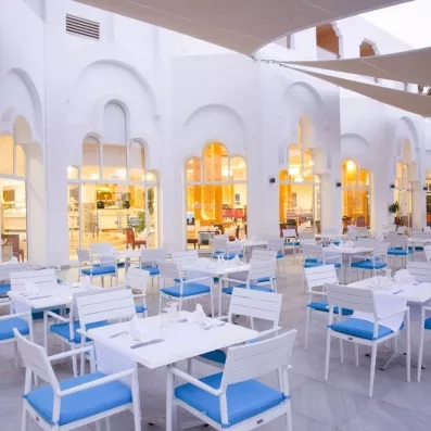 Terrasse extérieure lumineuse de l'hôtel Marhaba Palace à Monastir avec tables blanches et chaises bleues.