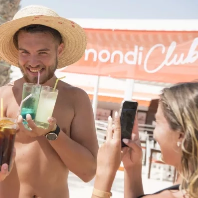 homme avec chapeau et cocktails et une femme qui le prend en photo avec son téléphone, bannière mondi club en arrière plan