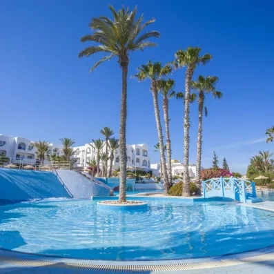 Grande piscine extérieure du Seabel Aladin Djerba avec petit pont bleu, toboggan et îlot de palmiers au centre.