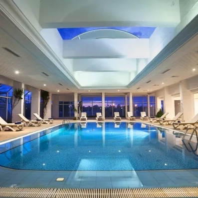 Piscine intérieure élégante avec chaises longues blanches, plafond voûté et grandes baies vitrées au coucher du soleil.”