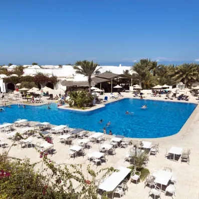 Piscine extérieure animée avec vacanciers, palmiers, parasols et bâtiments blancs à dômes.
