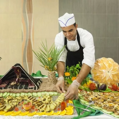 Chef en uniforme préparant un buffet décoratif avec fruits sculptés, légumes et présentation gastronomique.