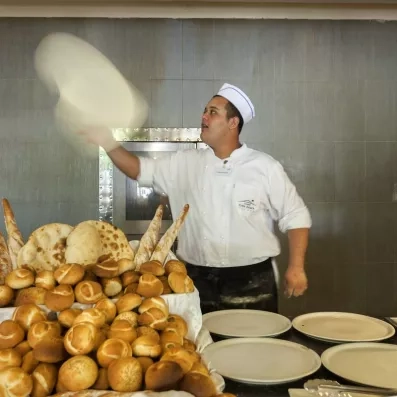 Boulanger en uniforme lançant une pâte à pizza devant un étal de pains variés.
