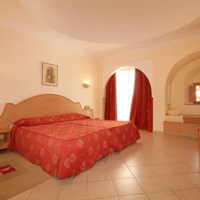 Chambre d'hôtel spacieuse à Zarzis avec un grand lit double couvert d'un couvre-lit rouge typique, une alcôve avec un lit d'appoint et une décoration aux influences tunisiennes.