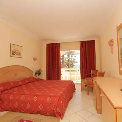 Chambre double lumineuse avec balcon privé offrant une vue sur les palmiers et la mer, équipée d'un mobilier en bois clair et de rideaux rouges chaleureux.