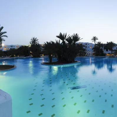 Vue nocturne de la piscine illuminée du Djerba Resort entourée de jardins exotiques et de palmiers.