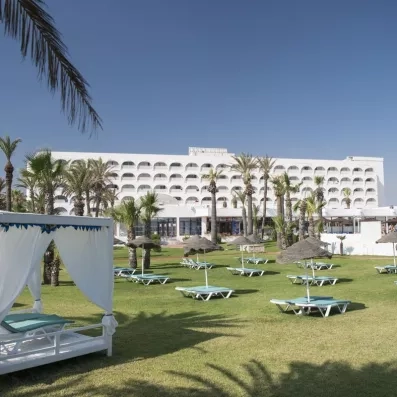 Jardin ensoleillé et façade de l'hôtel Mondi Club One Resort Jockey avec lits balinais et palmiers à Monastir, Tunisie.