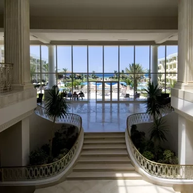 Hall de réception luxueux avec escalier monumental, grandes baies vitrées et vue sur jardin à Monastir.