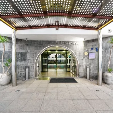 Une entrée de bâtiment moderne sous une pergola en bois, encadrée par des murs en pierre grise.