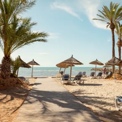 Ponton en bois menant à la plage de sable fin avec parasols en paille au Palm Azur Djerba.