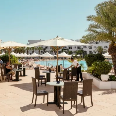 Terrasse ensoleillée de l'hôtel Palm Azur avec parasols, palmiers et vue sur la piscine bleue cristalline.