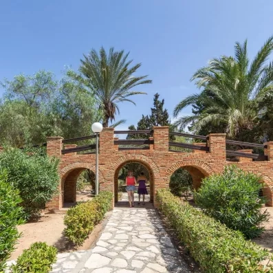 Promenade ensoleillée sous un pont en briques dans les jardins luxuriants du Marhaba Salem à Monastir.