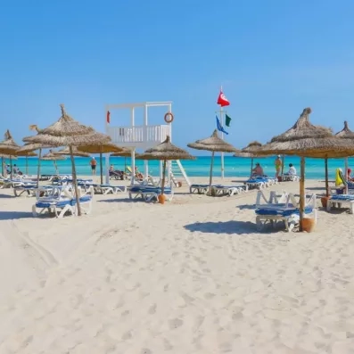 Plage de sable fin avec parasols en paille et mer turquoise au Royal Garden Palace de Djerba.