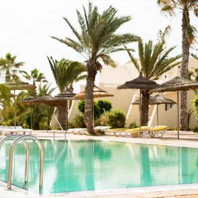 Espace détente avec piscine, parasols en paille et palmiers dans les jardins de l'hôtel à Djerba.