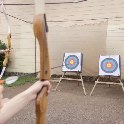 Une personne tient un arc en bois et vise deux cibles de tir à l'arc installées en extérieur.