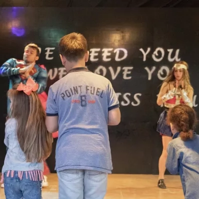 Des enfants assistent à un spectacle sur scène où des animateurs effectuent une chorégraphie.