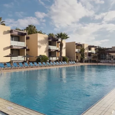 Une grande piscine bleue bordée de transats face à des bâtiments de vacances de style méditerranéen.