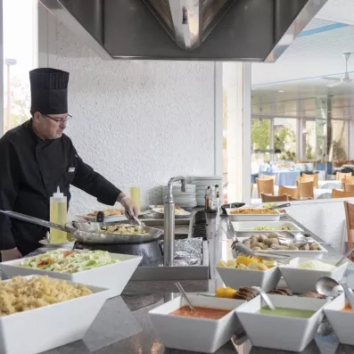 Un chef cuisinier prépare des plats devant un buffet varié dans une salle de restauration lumineuse.