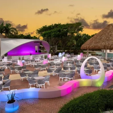 bar le soir, scène avec jeux de lumière pour les spectacles et les karaokés, bar sur la droite, tables et chaises pour s'asseoir et regarder, végétation au fond