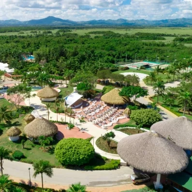 palmiers, végétation luxuriante, transats et parasols, bâtiments plein pied en paillote, au fond terrain de tennis