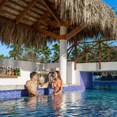 bar piscine, toit paillote, un homme et une femme en maillot de bain discutent dans la piscine en souriant et en buvant un cocktail, bouteilles dans le bar