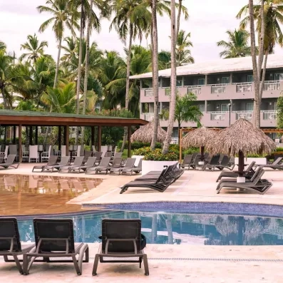 vue piscine, transats et parasols, façade de l'hôtel deux étages et balcons, nombreux palmiers végétation