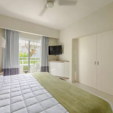 chambre familiale lit double avec porte fermée, écran accroché au mur, baie vitrée donnant sur un balcon avec table et chaises, vue sur le jardin et les palmiers