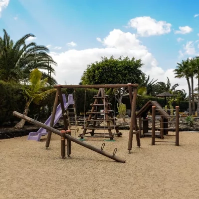 Une aire de jeux pour enfants en bois installée sur un sol souple, entourée de palmiers et de verdure.