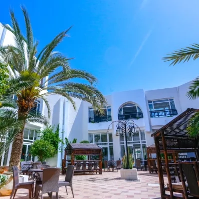 Cour intérieure ensoleillée de l'hôtel avec palmiers, mobilier de jardin et pavillons en bois.