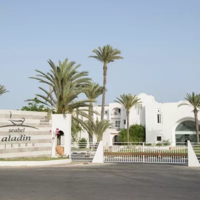 Entrée principale de l'hôtel Seabel Aladin Djerba avec enseigne, palmiers et architecture blanche traditionnelle sous un ciel bleu.