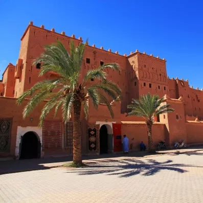 ouarzazate construction traditionnelle en brick rouge, deux palmiers devant la porte d'entrée et un ciel bleu