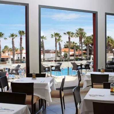 Une salle de restaurant lumineuse, offrant une vue panoramique sur les palmiers et la piscine.