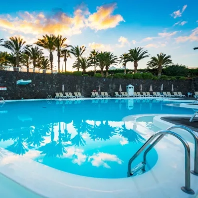 Une piscine calme au coucher du soleil, avec des palmiers et des transats en arrière-plan.