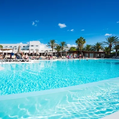 Une grande piscine d'hôtel s'étend sous un ciel bleu, bordée de palmiers.