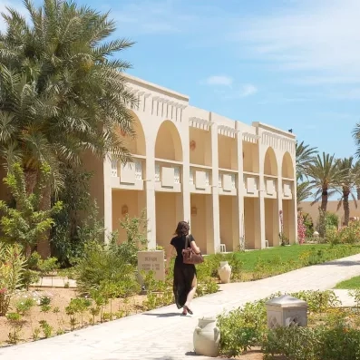 Façade de style architecture tunisienne traditionnelle avec arcades blanches, balcons fleuris et allée piétonne dans les jardins du complexe hôtelier.