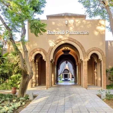 L'entrée hôtel avec arches et porte, au fond rideaux et fontaine, en haut lampe et toit, à côté arbres et plantes le long de l’allée