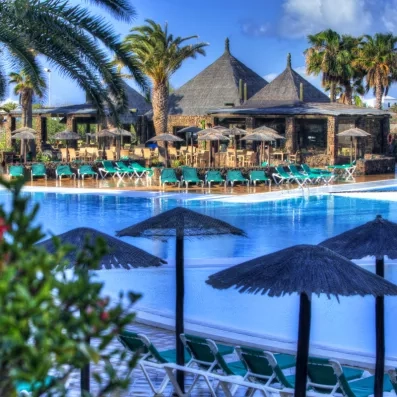 Une piscine bleu bordée de palmiers et de parasols sous un soleil éclatant.