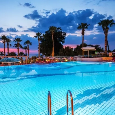 Une grande piscine extérieure, entourée de palmiers sous un ciel aux teintes rosées.