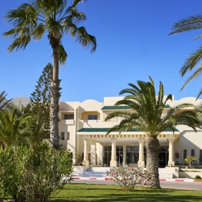 Façade blanche de l'hôtel Les Quatre Saisons Djerba entourée de palmiers sous un ciel bleu azur.