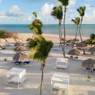 plage de sable, lits avec auvents blancs, transats bleus et parasols paillote, palmiers, eau de l'océan bleue, ciel bleu avec nuages