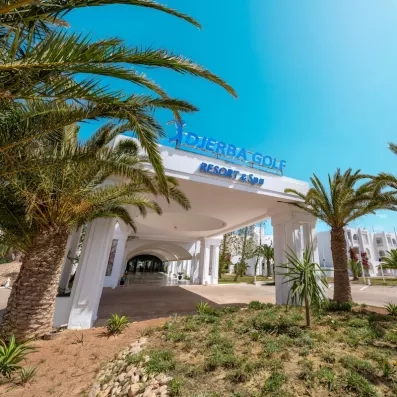 Façade blanche et entrée prestigieuse de l’hôtel Djerba Golf Resort & Spa sous un ciel bleu radieux.
