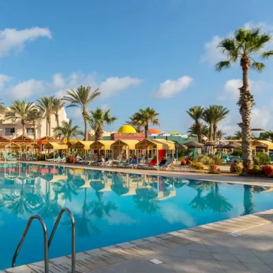 Grande piscine de resort entourée de cabanas jaunes, palmiers et transats, avec ciel bleu et ambiance tropicale relaxante.
