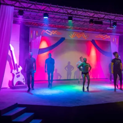 Groupe de danseurs sur scène éclairée par des lumières colorées, décor artistique avec guitare géante et drapés, ambiance de spectacle dynamique