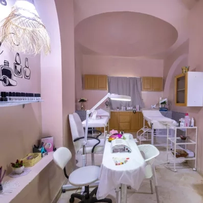 Intérieur d’un salon de beauté moderne avec tables de manucure, lampes UV, présentoirs de vernis et deux lits de soins dans une ambiance rose pastel.