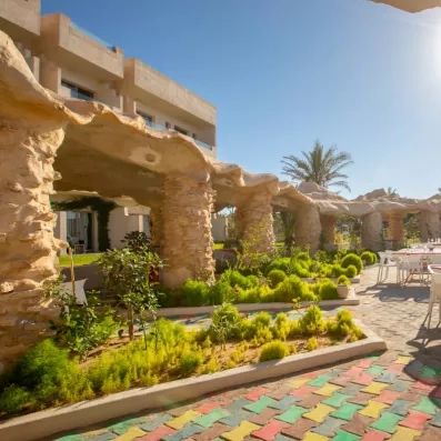 Passage couvert avec colonnes en pierre, toit ondulé et jardin paysager dans un resort moderne.