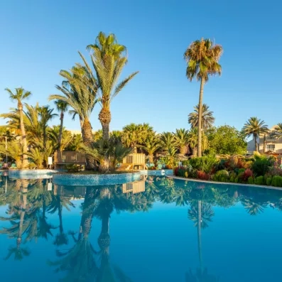 Piscine claire entourée de palmiers et structures en bois dans un cadre tropical ensoleillé.