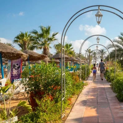 Allée pavée bordée de palmiers et lanternes décoratives menant à une plage tropicale.